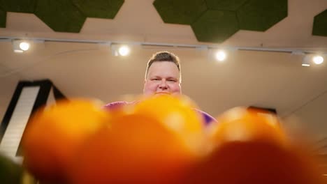 Shot-from-the-counter-of-a-happy-overweight-man-in-a-purple-T-shirt-choosing-oranges-from-the-counter
