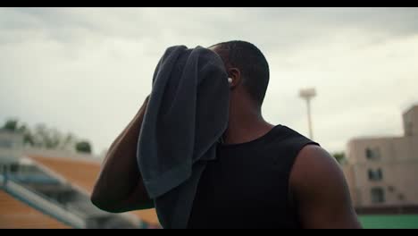 An-athlete-with-black-skin-in-a-black-T-shirt-wipes-his-face-with-a-towel-and-drinks-water-from-a-special-bottle-on-a-city-beach-against-a-gray-sky