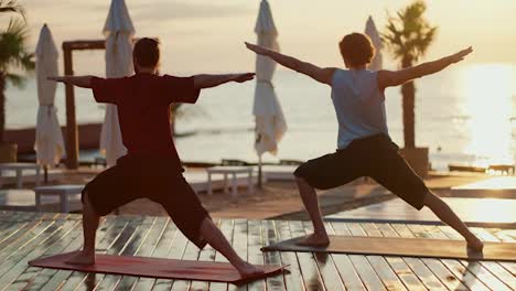 Meditation-of-two-guys-who-stand-on-a-wooden-floor-on-the-beach-and-stretch-their-arms-to-the-sides.-Yoga-and-Zen-style