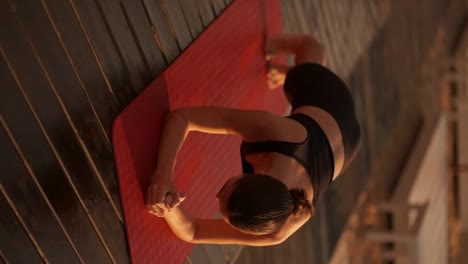 Vertical-video:-a-brunette-girl-in-a-black-tracksuit-does-a-plank-and-periodically-stands-on-her-elbows-on-a-sunny-beach-covered-with-black-boards-in-the-summer