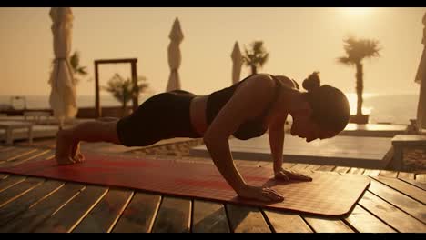 A-girl-in-a-sports-summer-uniform-with-tied-hair-does-push-ups-on-a-red-mat-on-a-sunny-beach-during-sunrise-in-summer