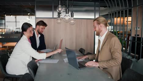 A-brunette-girl-in-a-white-suit-gives-a-blonde-man-in-a-light-brown-jacket-papers-for-work.-Paper-work-in-modern-office.-A-blond-man-with-a-beard-and-glasses-communicates-with-his-work-colleagues-at-a-table-in-the-office