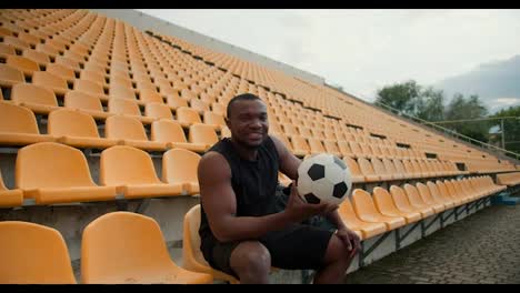 A-young-athlete-with-Black-skin-color-sits-in-a-black-sports-uniform-on-the-yellow-stands-of-the-stadium-and-holds-a-soccer-ball-in-his-hands.-Portrait-of-a-happy-and-successful-sportsman