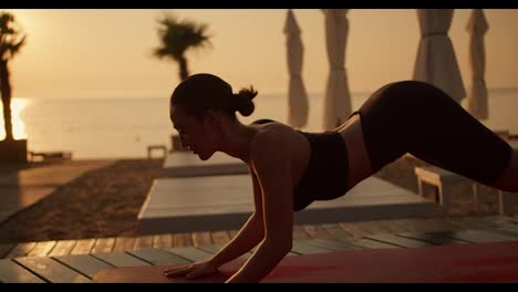 Shot-close-up-of-a-brunette-girl-with-tied-hair-in-a-black-sports-summer-uniform-doing-the-Cobra-exercise-and-doing-yoga-on-a-Sunny-beach-in-the-summer.-Golden-hour,-golden-sunrise