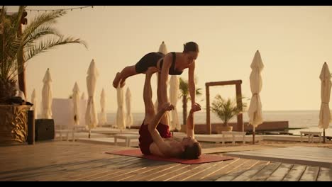 The-guy-holds-the-girl-up-with-his-legs,-the-couple-does-yoga-combined-with-acrobatics-on-a-red-carpet-on-a-sunny-beach-in-the