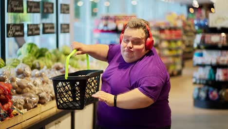 A-happy-and-cheerful-overweight-man-in-a-purple-T-shirt-and-red-headphones-walks-through-the-supermarket-and-dances-with-a-basket-in-his-hands,-choosing-fruits-for-himself.-A-man-tosses-an-apple-and-puts-it-in-his-basket