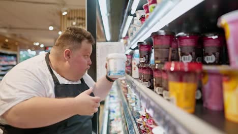 Serious-overweight-male-supermarket-worker-in-a-white-T-shirt-and-black-apron-lays-out-yogurt-and-sour-cream-on-the-counter-in-a