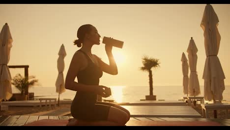 A-brunette-girl-in-a-black-sports-summer-suit-sits-on-her-knees-on-a-red-mat-and-drinks-water-from-a-sports-bottle-in-the-evening-during-a-golden-sunset-on-the-beach