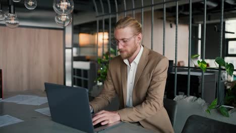 A-blond-guy-in-glasses-with-a-beard-in-a-light-brown-suit-works-at-a-laptop-and-then-looks-at-the-camera-and-smiles.-Portrait-of-a-modern-office-worker