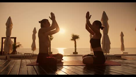 A-brunette-girl-in-a-black-top-and-a-guy-with-a-bare-torso-are-meditating-on-a-Red-Mat-on-a-sunny-beach-during-a-golden-dawn