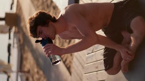 Close-up-shot-of-a-brunette-guy-with-a-naked-torso-drinking-water-from-a-special-sports-bottle-on-the-beach