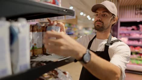 Serious-supermarket-worker-in-a-white-T-shirt-and-black-apron-lays-out-diet-vegan-milk-on-the-counter.-Working-with-milk-and-dairy-products-in-a-supermarket