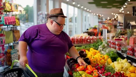 An-overweight-man-in-a-purple-T-shirt-with-glasses-and-a-brown-hat-walks-along-the-vegetable-counter-in-a-large-supermarket-and-chooses-tomatoes-and-zucchini.-A-review-of-a-trip-to-the-store-from-a-bright-man-and-from-a-bright-overweight-man