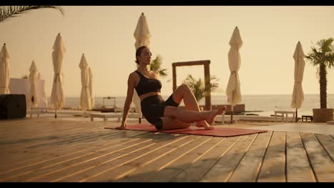 Ein-Mädchen-Massiert-Die-Rückseite-Ihres-Oberschenkels-Mit-Einem-Speziellen-Kegel-Auf-Einem-Roten-Teppich-An-Einem-Sonnigen-Strand,-Der-Mit-Brettern-Bedeckt-Ist.-Yoga
