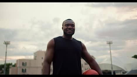 Ein-Schwarzer-Mann-In-Einem-Schwarzen-T-Shirt-Mit-Einem-Basketball-In-Der-Hand-Posiert-Und-Blickt-Vor-Einem-Grauen-Himmel-In-Die-Kamera