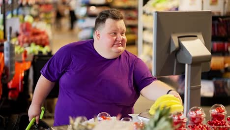A-happy-overweight-man-in-a-purple-T-shirt-and-short-hair-puts-bananas-on-special-scales-in-the-supermarket-to-get-a-price-tag-on-them-and-find-out-the-weight-of-the-fruit