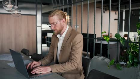A-blond-guy-with-glasses-in-light-brown-is-at-a-laptop-and-looks-at-the-camera.-Portrait-of-a-young-male-businessman-at-his-workplace-in-a-modern-office