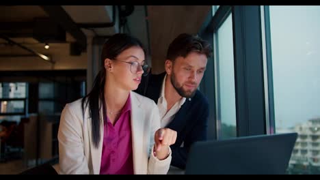 A-brunette-girl-in-a-white-jacket-and-pink-shirt-with-round-glasses-sits-with-a-guy-in-a-blue-jacket-and-works-on-a-laptop-near-a-panoramic-window-in-the-office