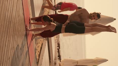 Vertical-video:-a-brunette-guy-in-red-pants-with-a-bare-torso-helps-a-girl-in-a-black-sports-summer-uniform-to-stand-on-her-hands-with-her-head-down-on-a-sunny-beach-on-a-red-carpet