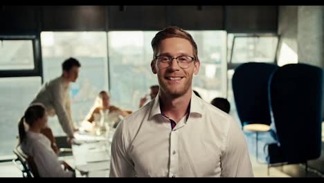 A-blond-guy-in-glasses-and-a-white-shirt-poses-in-the-office-against-the-background-of-his-gollegs-who-work-at-a-table-in-an-office-with-panoramic-windows
