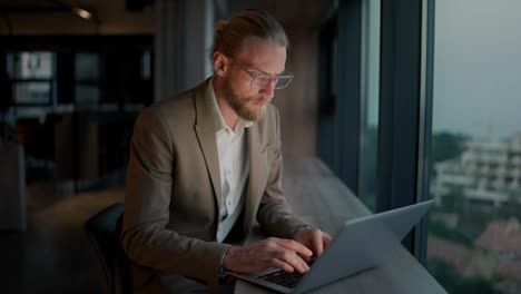 A-blond-guy-in-glasses-and-a-light-brown-suit-notices-an-error-on-the-laptop-screen-and-thinks-about-its-solution-in-a-modern-office-near-the-window