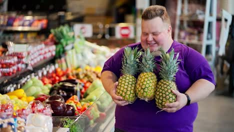 Portrait-of-a-happy-overweight-man-wearing-a-purple-T-shirt-and-a-short-haircut-who-holds-three-pineapples-in-his-hands-smiles-and-looks-at-the-camera-in-a-large-supermarket-near-a-counter-with-vegetables