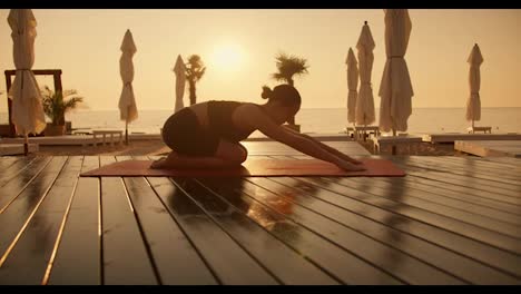 A-brunette-girl-with-tied-hair-in-a-black-sports-summer-uniform-does-yoga-on-red-mats-on-the-seashore-with-a-yellow-sunrise.-The-girl-takes-Half-Tortoise-Pose-during-a-yoga-class