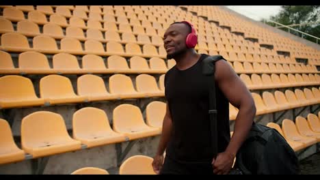 Portrait-of-an-athlete-with-Black-skin-in-a-black-jersey-and-red-wireless-headphones,-who-walks-through-the-stadium-with-yellow-chairs-in-the-stands,-and-looking-for-his-place