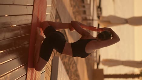 Vertical-video:-A-brunette-girl-with-tied-hair-in-a-black-sports-summer-uniform-takes-a-special-stance-during-a-yoga-class-on-a-special-red-mat-on-a-sunny-beach-during-a-golden-sunset-in-summer