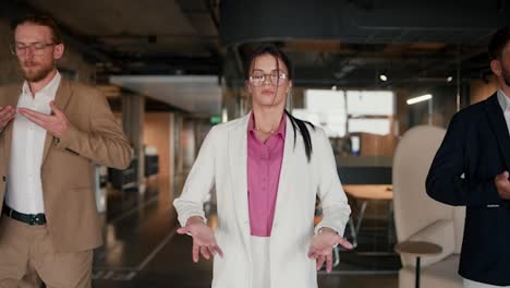 Close-up-shot-of-a-brunette-girl-in-round-glasses-in-a-white-suit-with-a-pink-shirt-doing-breathing-techniques-with-her-colleagues-during-a-break-at-work-in-a-modern-office