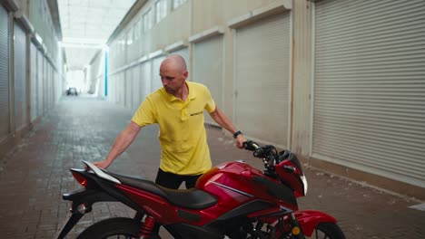 Side-view.-A-bald-man-driving-instructor-in-a-yellow-T-shirt-drives-a-red-moped-into-the-garage.-End-of-the-working-day-at-the-motorcycle-school