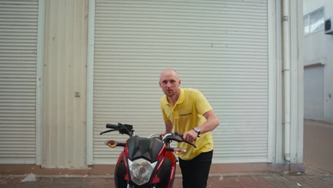 A-bald-man-driving-instructor-in-a-yellow-T-shirt-drives-a-red-moped-into-the-garage