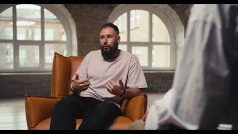 An-excited-man-with-a-beard-in-a-pink-shirt-talks-about-his-problems-and-questions-to-a-psychologist.-A-young-man-sits-on-a-brown-chair-and-communicates-with-a-psychologist-in-a-special-room-made-of-bricks