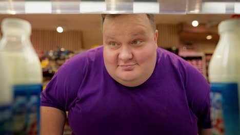Happy-overweight-man-in-purple-t-shirt-raises-eyebrow-and-takes-dairy-product-from-shelf-in-supermarket.-Selection-of-milk-in-a-supermarket