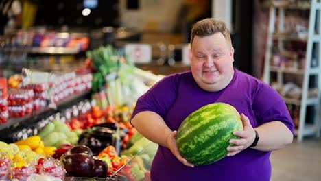 Portrait-of-a-happy-overweight-man-in-a-purple-T-shirt-who-smiles-and-looks-at-the-camera-and-holds-a-large-and-green-watermelon-in-his-hands-in-a-supermarket-near-the-counter-with-vegetables
