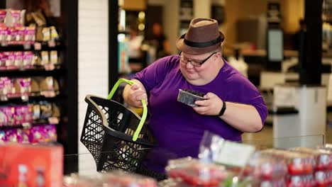 An-overweight-man-in-a-purple-T-shirt-and-a-brown-hat-smells-berries-in-a-supermarket-and-puts-them-in-his-basket.-Walking-with-a-bright-personality,-an-overweight-man-chooses-berries-for-himself