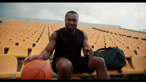 Portrait-of-a-Happy-sportsman-with-Black-skin-color-in-a-black-jersey-who-hits-a-basketball-from-the-ground-and-looks-at-the-camera.-Happy-young-sportsman-in-the-yellow-stands-of-the-stadium
