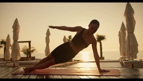A-girl-in-a-black-sports-summer-uniform-does-a-plank-exercise-on-one-arm-on-a-red-mat-on-the-beach-on-the-shore-of-the-sunny-sea