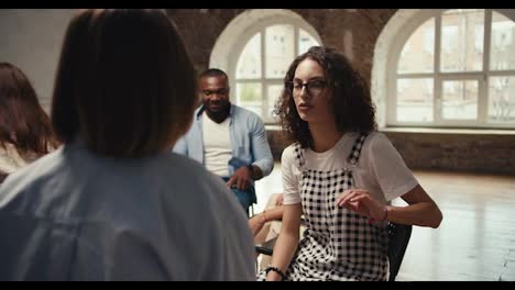 A-brunette-girl-with-curly-hair-in-glasses-and-a-checkered-jumpsuit-communicates-in-pairs-during-group-therapy-in-a-brown-hall