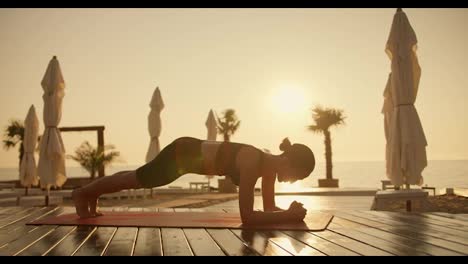 A-brunette-girl-in-a-black-top-does-push-ups-on-her-elbows-on-a-Red-mat-on-a-Sunny-beach-which-is-covered-with-boards.-Sports