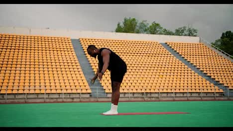 Black-man-in-black-sportswear-doing-yoga-in-a-green-stadium-with-yellow-chairs-in-the-stands