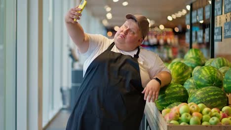 A-funny-and-happy-overweight-male-supermarket-worker-in-a-white-T-shirt-and-a-black-apron-in-a-gray-cap-takes-a-selfie-using-a-yellow-phone-against-the-background-of-watermelons-in-the-supermarket