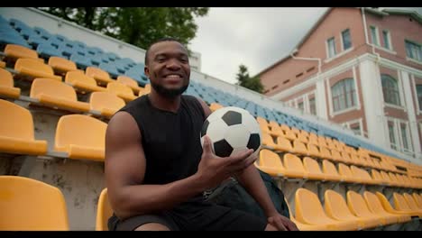 A-Black-skinned-athlete-in-a-black-sports-summer-uniform-poses-on-the-yellow-stands-of-the-stadium-with-a-soccer-ball-in-his-hands