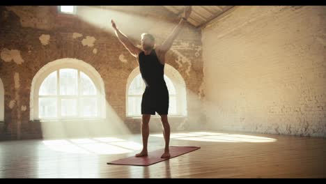 A-blond-man-in-black-clothes-does-exercises-on-a-special-mat-in-a-spacious-sunny-room-with-brick-walls