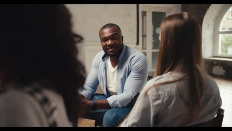 A-man-with-black-skin-in-a-blue-shirt-communicates-with-a-blonde-girl-at-group-therapy-in-a-white-room.-Communication-in-couples-in-group-therapy