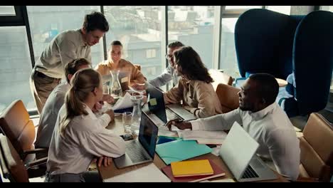 A-group-of-office-workers-sit-at-a-table-and-show-the-manager-guy-in-a-white-shirt-their-projects-on-paper.-Team-work-at-a-table-in-an-office-with-panoramic-windows