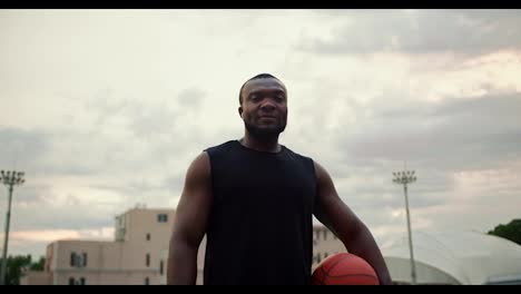 Serious-male-athlete-with-Black-skin-color-in-a-black-T-shirt-posing-at-the-city-stadium-with-a-basketball-in-his-hands-against-a-gray-sky