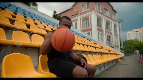A-Black-skinned-athlete-in-a-sports-summer-uniform-sits-on-the-stands-of-a-yellow-stadium,-hits-a-basketball-ball-on-the-floor-and-looks-at-the-camera