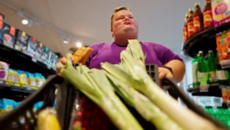 View-from-the-cart-side:-A-happy-overweight-man-wearing-a-purple-T-shirt-walks-through-the-supermarket,-looks-at-the-products-and-eats-a-croissant.-Shallots-are-already-in-the-man's-cart