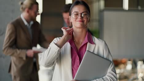 Portrait-of-a-brunette-girl-in-round-glasses-in-a-white-jacket-and-pink-shirt-who-stands-with-a-laptop-and-adjusts-her-glasses-against-of-a-modern-office-and-her-colleagues